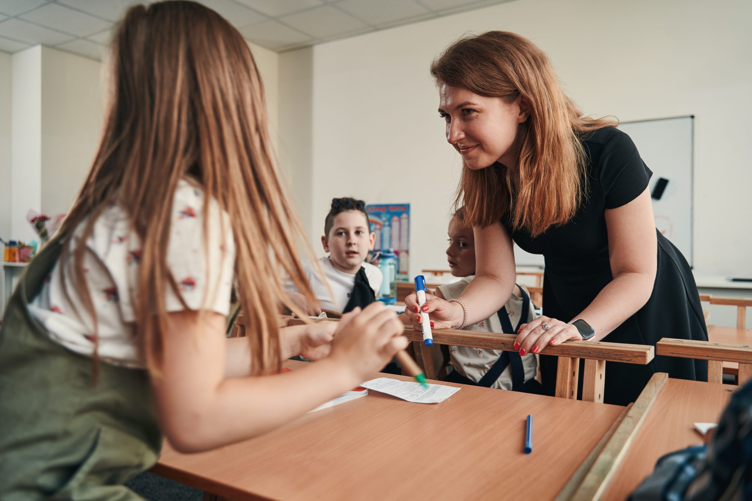 Friendly smiling teacher holding a marker while leaning towards the desk and looking at the pupil